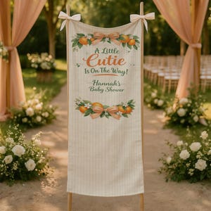 May include: A beige banner with the text "A Little Cutie Is On The Way! Hannah's Baby Shower." The banner is decorated with orange illustrations, bows, and floral accents. The banner is held up by wooden poles and tied with white bows.