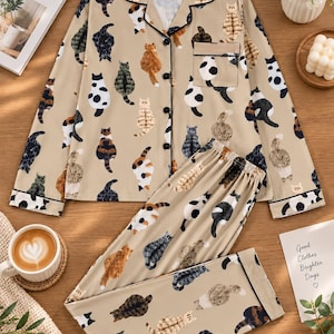 May include: A beige pajama set featuring a long-sleeved button-down shirt with a notched collar and a chest pocket, paired with matching long pants. The entire set is covered in a repeating pattern of various cat breeds in black, white, and brown.