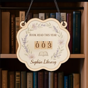 Puede incluir: Letrero de madera con diseño floral y el texto "Book Read This Year". El letrero tiene un contador que muestra "003" y las palabras "Sophia Library". Cuelga de una cadena frente a una estantería.