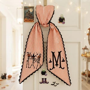 May include: A long, orange and white striped fabric door hanger with a scalloped black edge. One side features embroidered skeletons wearing pumpkin heads, while the other side displays a large black letter M flanked by two small witch hats.