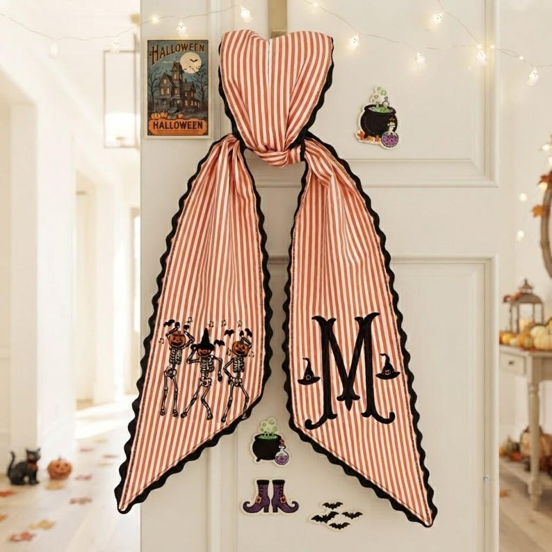 May include: A long, orange and white striped fabric door hanger with a scalloped black edge. One side features embroidered skeletons wearing pumpkin heads, while the other side displays a large black letter M flanked by two small witch hats.