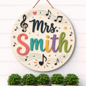 May include: A round wooden sign featuring the text Mrs. Smith in colorful raised lettering. The background is cream with black musical staves, black musical notes, and small colorful hearts and stars. A rope hanger is attached to the top.
