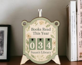 May include: A wooden book counter with the text "Books Read This Year" and the number "034". The counter has a floral design and the text "Susan's Library". The counter is on a shelf with books.