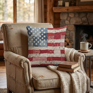 May include: A decorative pillow with a patchwork design resembling the American flag, featuring red and white stripes and a blue field with white stars. The pillow is placed on a beige armchair, creating a cozy, patriotic theme.
