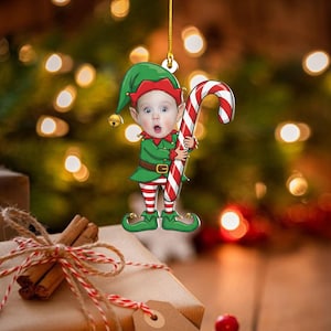 Puede incluir: Adorno navideño que representa a un niño disfrazado de elfo, sosteniendo un bastón de caramelo. El elfo viste un traje verde con un gorro rojo y medias a rayas. Se adjunta un hilo dorado para colgar.