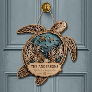 Op de afbeelding: Een gelaagde houten wandhanger in de vorm van een zeeschildpad met een gedetailleerde onderwater koraalrifscène in het schild. Het object bevat de tekst THE ANDERSONS en Together We Navigate Life en hangt aan een touw van jute.