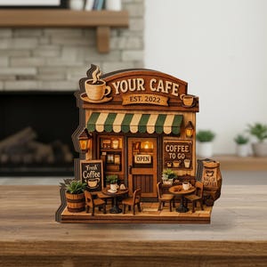 May include: A wooden model of a cafe with the text "YOUR CAFE EST. 2022". The cafe features a green and white striped awning, a sign that says "Fresh Coffee", and small tables and chairs. There is a sign that says "Coffee To Go".