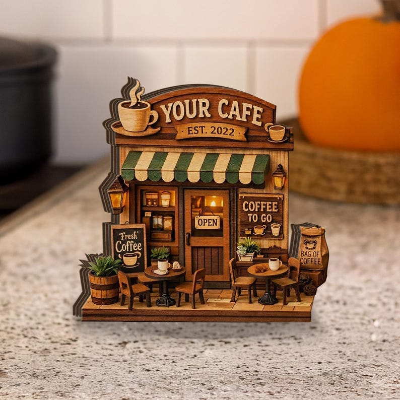 May include: A detailed wooden model of a cafe, featuring the text "YOUR CAFE EST. 2022". It has a green and white striped awning, signs for "Fresh Coffee" and "Coffee To Go", and miniature tables and chairs. The model is intricately designed.