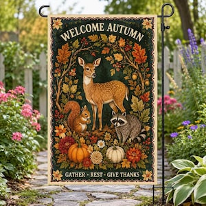 以下が含まれることがあります： 上部に「WELCOME AUTUMN」、下部に「GATHER REST GIVE THANKS」の文字が記された長方形のガーデンフラッグ。秋の葉、どんぐり、オレンジと緑のカボチャに囲まれた鹿、リス、アライグマが描かれています。
