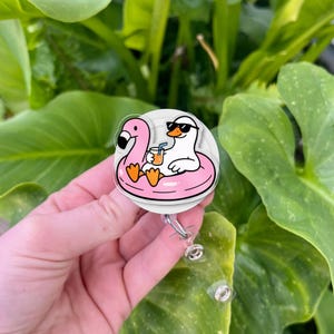 Puede incluir: Un portacredenciales circular con un pato blanco de dibujos animados que lleva gafas de sol negras mientras flota en un flotador rosa con forma de flamenco. El pato sostiene una bebida naranja con una pajita azul y tiene una correa transparente.