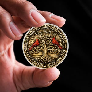 Peut inclure: Une pièce de monnaie ronde dorée avec un arbre et deux oiseaux cardinaux rouges. La pièce porte l'inscription "Always With Me". La pièce est tenue entre deux doigts sur un fond noir.