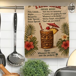Könnte beinhalten: Ein beiges Küchentuch mit Waffelstruktur, das ein Tiki-Bar-Getränkemenü mit Text, eine Tiki-Becher-Illustration mit Strohhalm, Kirsche und Cocktailschirmchen zeigt, umgeben von roten und weißen Hibiskusblüten und grünen Palmblättern.