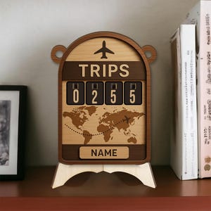 Könnte beinhalten: Reisezähler aus Holz mit dem Wort "TRIPS" über einer Digitalanzeige mit der Anzeige "0255". Unter der Anzeige ist eine Weltkarte eingeätzt, mit einer gestrichelten Linie und einer Flugzeuggrafik. Das Wort "NAME" steht unten.