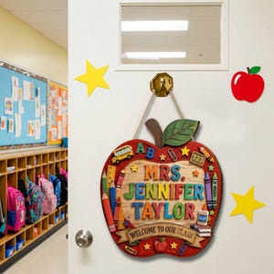 May include: A wooden apple-shaped classroom door sign featuring the text MRS. JENNIFER TAYLOR and WELCOME TO OUR CLASS. The sign is decorated with colorful illustrations of pencils, crayons, a school bus, and a glue bottle, hanging by a rope.