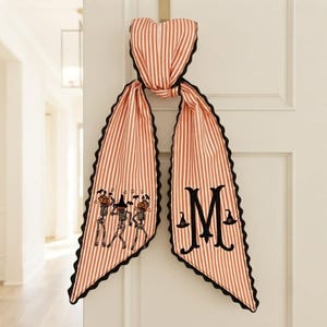 May include: A long, orange and white striped fabric door hanger with scalloped black edges. One side features embroidered skeletons with pumpkin heads, while the other side displays a large black letter M flanked by two small witch hats.