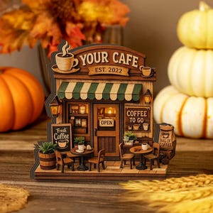 May include: A wooden model of a cafe with the text "YOUR CAFE EST. 2022". The cafe features a green and white striped awning, small tables, chairs, and a sign that says "Fresh Coffee". There is a bag of coffee and a sign that says "Coffee To Go".