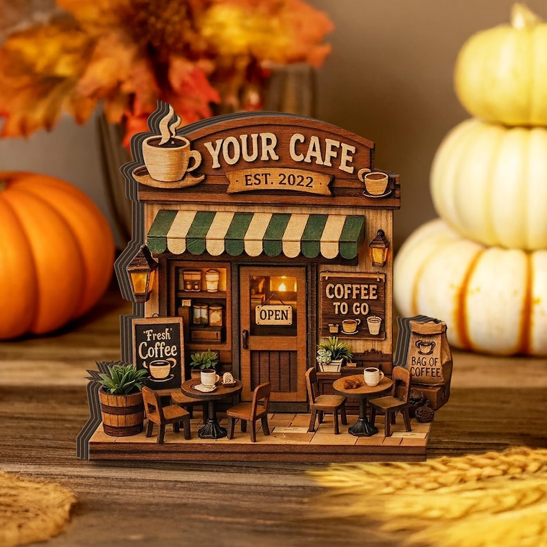 May include: A wooden model of a cafe with the text "YOUR CAFE EST. 2022". The cafe features a green and white striped awning, small tables, chairs, and a sign that says "Fresh Coffee". There is a bag of coffee and a sign that says "Coffee To Go".