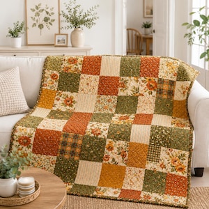 Könnte beinhalten: Eine Patchwork-Decke mit einem Raster aus quadratischen Stoffblöcken in den Farben gebranntes Orange, Olivgrün, Senfgelb und Creme. Das Design umfasst verschiedene Blumenmuster, kleine Streublumen-Prints und feine vertikale Streifen.