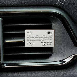 May include: A rectangular, silver-colored car vent clip with the word "Daddy" at the top. The clip has a heartfelt message, angel wings, and two hearts. The message is signed "Katie - May 2023".