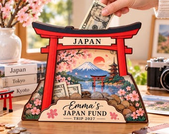 Può includere: Un salvadanaio in legno a forma di portale torii rosso con pannello frontale trasparente. L'interno presenta una scena stampata del Monte Fuji, fiori di ciliegio e una pagoda. Il testo riporta JAPAN e Emma's JAPAN FUND TRIP 2027.
