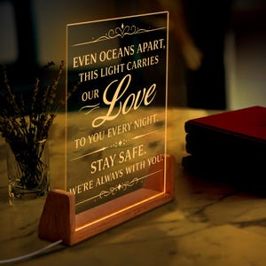 May include: A clear acrylic LED light with the words "EVEN OCEANS APART, THIS LIGHT CARRIES OUR Love TO YOU EVERY NIGHT. STAY SAFE. WE'RE ALWAYS WITH YOU." The light sits on a wooden base and is illuminated with a warm glow.