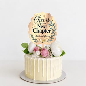 以下が含まれることがあります： Cheers to the Next Chapter retired and glowingという文字、本のアイコン、青い葉のイラストが描かれた金色のスカラップ型ケーキトッパー。白い花とピンクの花、白いアイシングの垂れ飾りで装飾された白いケーキの上に飾られている。