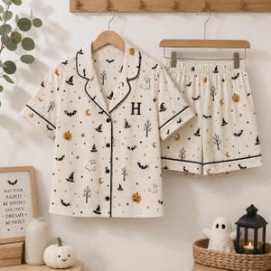 Puede incluir: Un conjunto de pijama color crema que incluye una camisa de manga corta con botones y pantalones cortos a juego. La tela presenta un estampado de pequeñas calabazas naranjas, murciélagos negros, sombreros de bruja, fantasmas, estrellas y árboles.