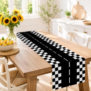 May include: A rectangular table runner featuring a black and white checkered pattern along the edges and a central black strip with a white dashed line, resembling a race track, laid flat across a wooden dining table surface.