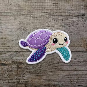 Può includere: Una toppa ricamata a forma di tartaruga marina con guscio viola, testa color crema con grandi occhi neri e guance rosa, e pinne con motivi viola e turchese, il tutto contornato da un bordo bianco spesso.