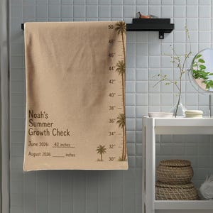 Könnte beinhalten: Ein beigefarbenes Wachstumsmess-Handtuch mit einem Palmenmotiv am rechten Rand und Messmarkierungen von 30 bis 50 Zoll. Der Text lautet Noah's Summer Growth Check, June 2026: 42 inches und August 2026: inches mit einer leeren Linie.