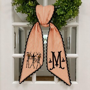 May include: A decorative fabric bow with orange and white vertical stripes and black scalloped edges. One tail features embroidered skeletons with pumpkin heads and bats, while the other tail displays a large black letter M with witch hats.