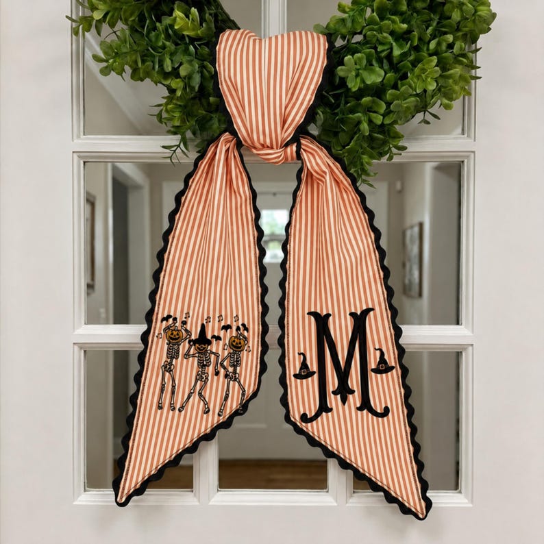 May include: A decorative fabric bow with orange and white vertical stripes and black scalloped edges. One tail features embroidered skeletons with pumpkin heads and bats, while the other tail displays a large black letter M with witch hats.