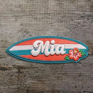 Può includere: Una toppa ricamata a forma di tavola da surf con strisce orizzontali color corallo, bianco e turchese. Il nome Mia è ricamato in grandi lettere corsive bianche al centro, con un fiore di ibisco rosa e foglie verdi sul lato destro.