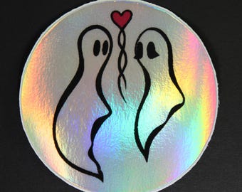 May include: A round, holographic sticker featuring two ghost figures connected by a red heart. The ghosts are outlined in black, with simple oval eyes. The sticker has a rainbow sheen.