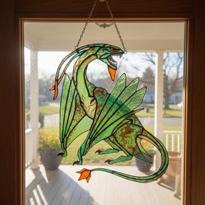 May include: Stained glass dragon window hanging. The dragon features a green body with brown accents, orange accents, and large green wings. The dragon is suspended by a chain and is displayed in a window. The dragon is a fantasy-themed decoration.