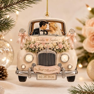 May include: A cream-colored vintage car ornament featuring a bride and groom figurine inside. The car is decorated with pink ribbons, white and pink flowers, and a banner reading Charlie and Sophia. A Just Married sign and 02.28.2026 license plate.