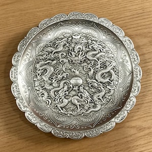 May include: A silver-colored decorative plate with an intricate dragon design. The plate features multiple dragons in a circular pattern, with detailed scales and features. The plate has a scalloped edge and is likely made of metal.