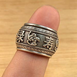 Può includere: Un anello color argento con caratteri cinesi incisi e motivi decorativi. L'anello ha una forma cilindrica e viene indossato su un dito. Il design dell'anello include dettagli intricati e una superficie strutturata.