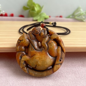 May include: A carved pendant featuring a monkey riding a horse, in shades of brown and tan. The pendant is attached to a black cord necklace with a red bead accent. The design is detailed and the pendant has a smooth finish.