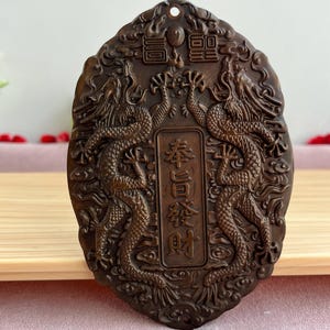 Op de afbeelding: Een bruine, ovale hanger met ingewikkelde drakenbewerkingen. Twee draken staan tegenover elkaar en omlijsten een centraal paneel met Chinese karakters. De hanger heeft een gat aan de bovenkant om op te hangen.