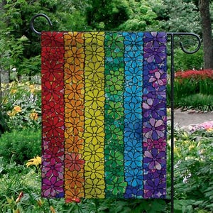 May include: A rectangular garden flag featuring a rainbow floral design. The flag displays vertical stripes of red, orange, yellow, green, blue, and purple, each filled with daisy-like flowers. The flag is hanging on a black metal stand in a garden setting.