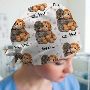 May include: White surgical scrub cap with a repeating pattern of hugging cartoon monkeys and pink hearts. The words "stay kind" are printed on the cap. The cap has a tie closure.