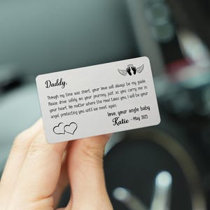 Peut inclure: Une carte en métal argenté avec le texte "Daddy." et un message sincère. La carte comprend des ailes d'ange et des empreintes de pas, ainsi que le nom "Katie - May 2025."