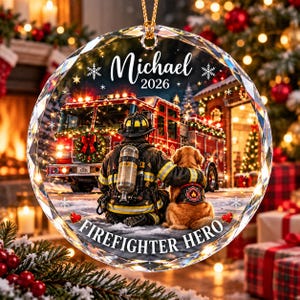 Könnte beinhalten: Ein kreisförmiges Glasornament mit einem Feuerwehrmann in Uniform, der mit einem Golden Retriever vor einem Feuerwehrauto sitzt. Der Text Michael 2026 steht oben und der Text FIREFIGHTER HERO ist unten mit Schneeflocken-Akzenten aufgedruckt.