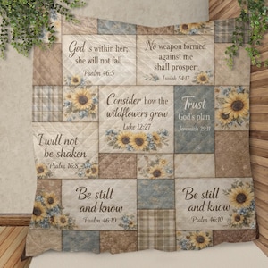 Op de afbeelding: Een gewatteerde deken met een patchwork-ontwerp met bloemen- en schrifttekst. De deken heeft een neutraal kleurenpalet met vierkanten in beige, blauw en ruitpatronen. Zonnebloemillustraties en inspirerende citaten zijn overal in het ontwerp te zien.