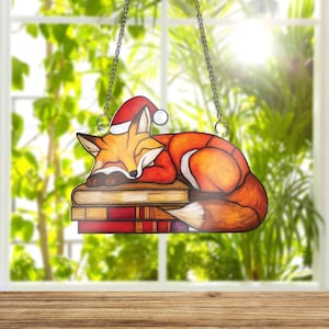 May include: A stained glass window decoration featuring a sleeping fox wearing a Santa hat, resting on a stack of books. The fox is orange, white, and brown, with a red hat. The books are various shades of red and brown. The decoration hangs from a silver chain.