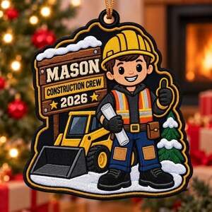 Peut inclure: Une décoration ornementale représentant un ouvrier de chantier en dessin animé avec un casque jaune, un gilet de sécurité orange et un pantalon bleu, tenant un plan. Un panneau en bois indique MASON CONSTRUCTION CREW 2026. Un chargeur jaune et un sapin enneigé sont visibles.