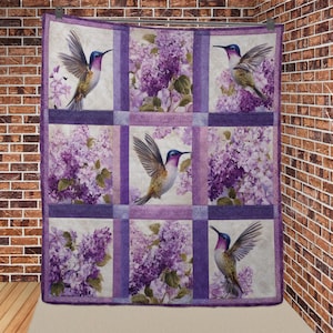 May include: A quilted throw blanket featuring a repeating pattern of hummingbirds and lilac flowers. The design incorporates squares of lilac and white, with the birds in flight. The color palette includes shades of purple, lavender, and green.