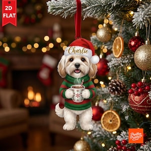 Può includere: Una decorazione natalizia piatta da appendere raffigurante un cane dal pelo chiaro e soffice, che indossa un maglione a righe rosse e verdi e un cappello da Babbo Natale rosso con il nome Charlie in oro. Il cane tiene in mano una tazza bianca.
