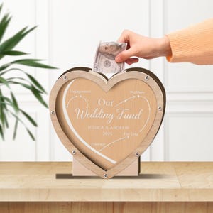 Può includere: Un salvadanaio a forma di cuore in legno con la scritta "Our Wedding Fund" e i nomi "Jessica & Andrew 2025". Una mano sta inserendo una banconota da 100 dollari nella fessura superiore. La scatola è di colore marrone chiaro.